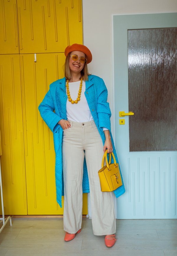 Naine oranži bareti, kollaste päikeseprillide, sinise mantli, valge särgi, beežide pükste, oranžide kingade ja kollase kaelakeega, hoides kollast käekotti, seisab siseruumides.