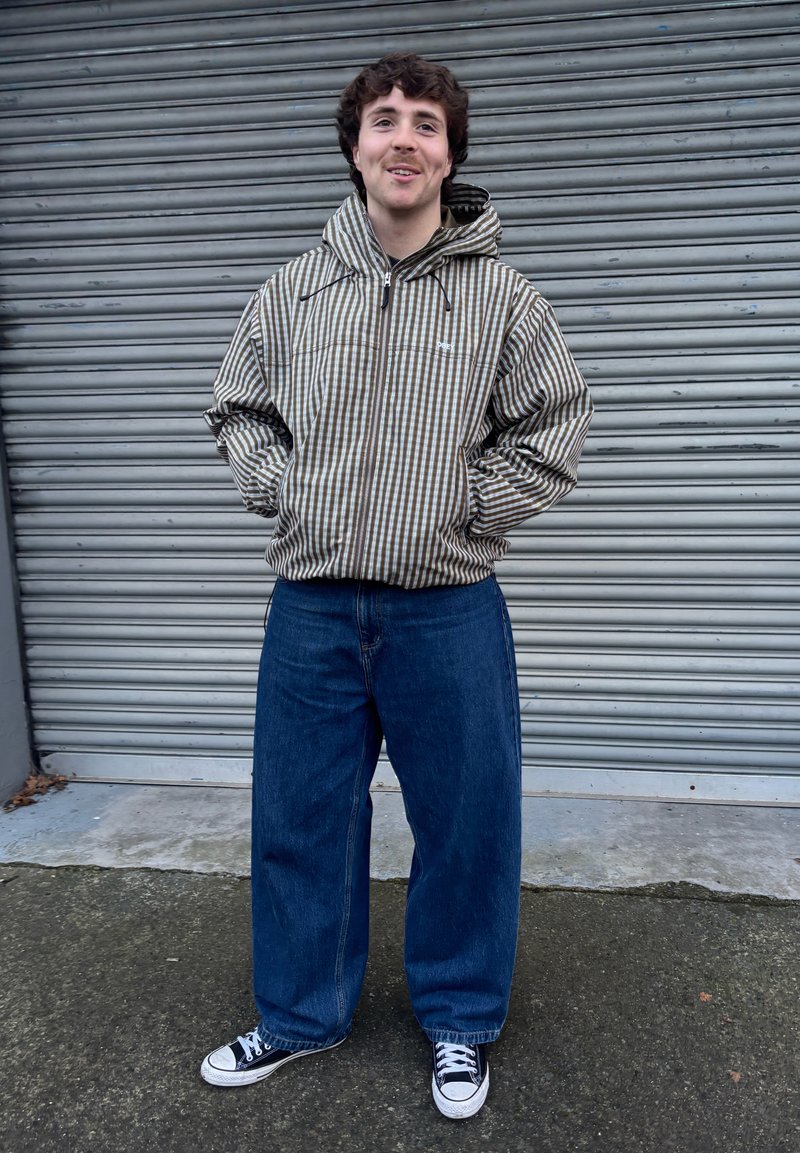 Giovane sorridente con le mani nelle tasche, indossa una giacca con cappuccio a righe, jeans larghi blu e sneakers bianche e nere, in piedi all'aperto.