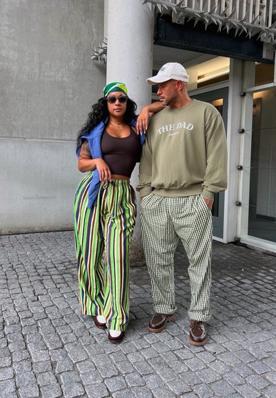 Dos personas están juntas sobre adoquines, vistiendo pantalones a rayas brillantes, un suéter azul y una sudadera verde oliva con texto.