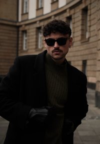 Homme aux cheveux bouclés portant des lunettes de soleil noires, un manteau sombre, des gants en cuir et un pull vert olive, debout dans une rue de la ville bordée de bâtiments en pierre.