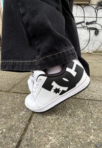 Baskets blanches avec un logo DC noir et un détail étoile, présentant une tige en cuir texturé, un bout rond et des lacets blancs sur une semelle noire.