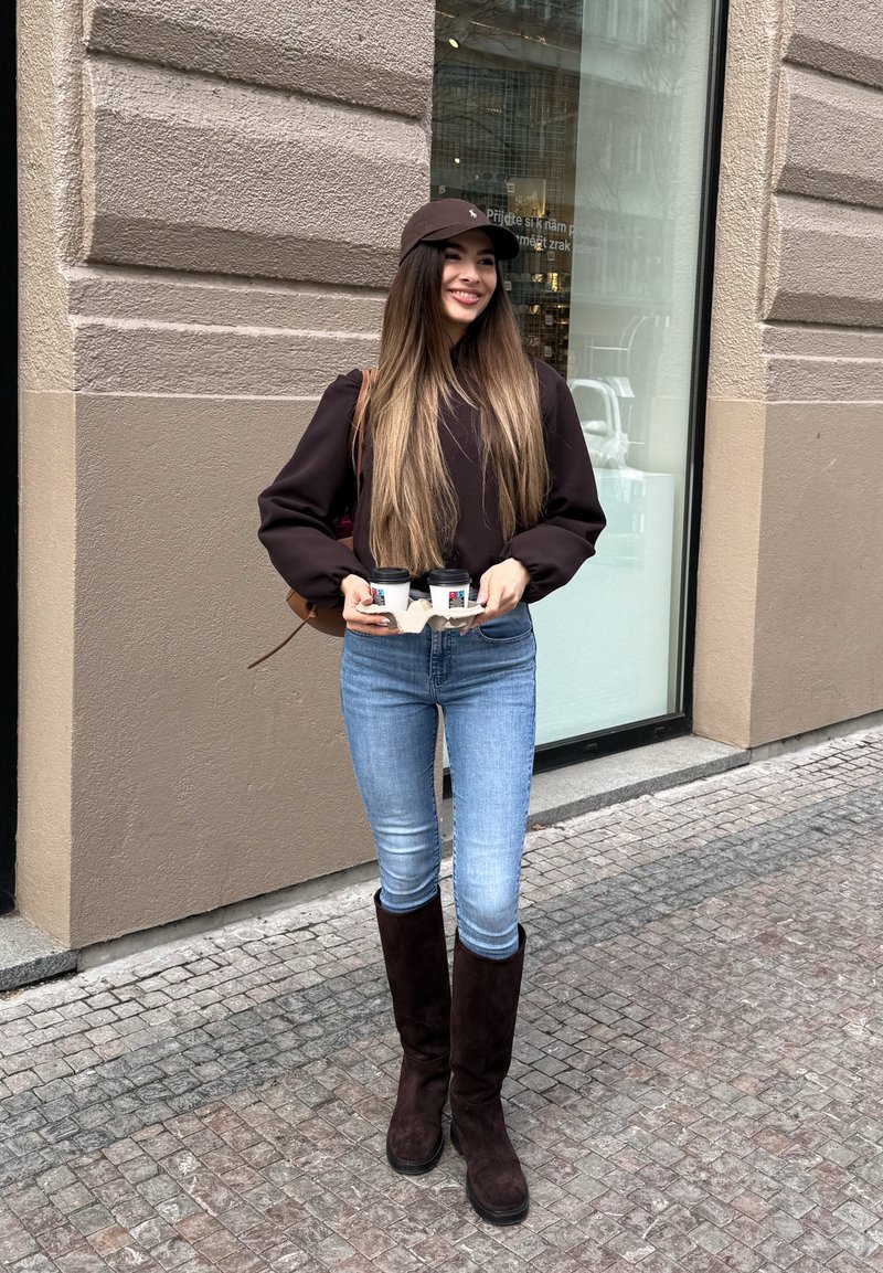 Jeune femme aux longs cheveux, casquette marron et bottes, tenant deux gobelets de café à emporter, debout dans une rue pavée près d'un immeuble.