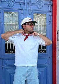 Biela tričko s krátkym rukávom v kombinácii s svetlomodrými pruhovanými šortkami. Okolo krku je viazaná červená bandana. Osoba má na sebe bielu čapicu a okuliare.
