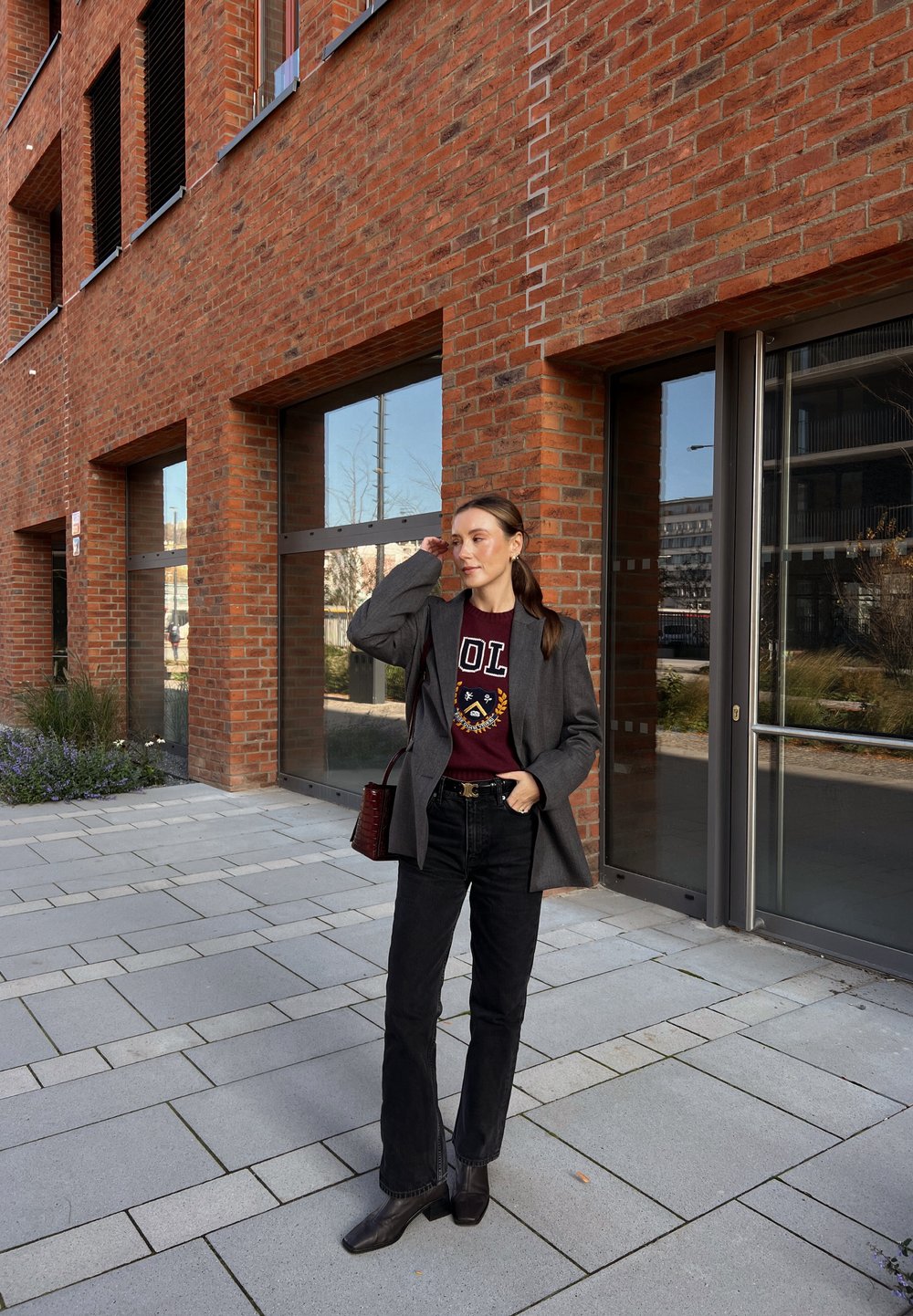 Blazer cinza oversized sobre um sweatshirt bordô com um logo, jeans pretos flare e botas de chelsea pretas de couro; a bolsa é de um vermelho escuro.