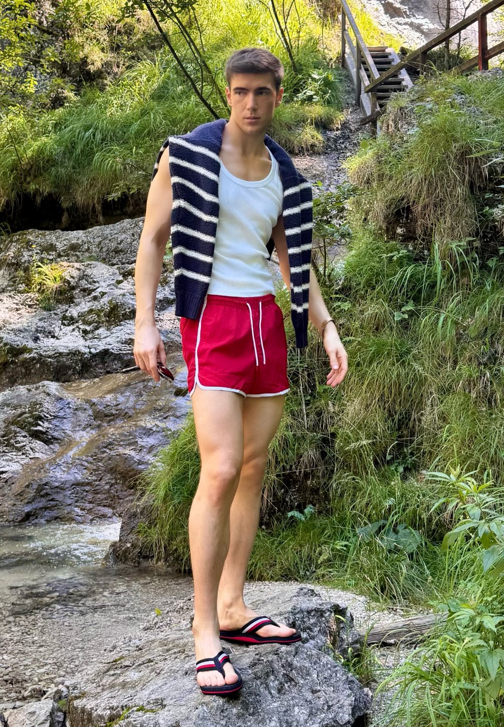 Figura masculina com uma camiseta branca sem mangas, calções vermelhos e chinelos pretos; suéter riscado em azul-marinho pousado sobre os ombros; terreno rochoso e vegetação.