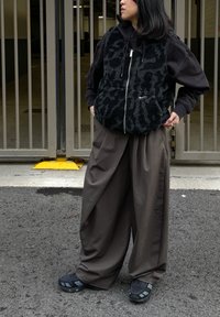 Sort fleecevest med tekstureret design, kombineret med en sort hættetrøje, løstsiddende brune bukser og sorte sko med fremtrædende detaljer.