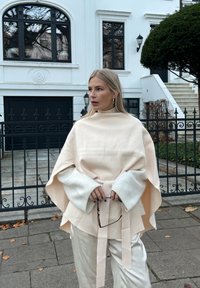 Svijetlo bež pončo s visokim ovratnikom i širokim rukavima, slojevit preko kremaste majice. Uparen s bijelim hlačama i drži naočale.