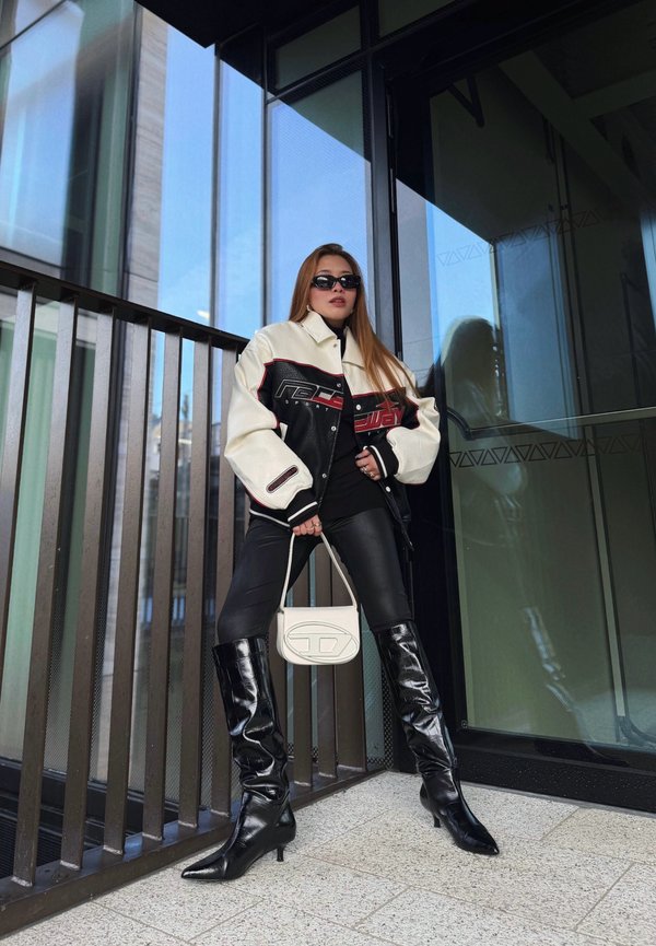 Femme portant des lunettes de soleil noires, une veste noire et blanche surdimensionnée, un pantalon noir et des bottes noires montant jusqu'aux genoux, se tient près d'une porte en verre en tenant un petit sac à main blanc.