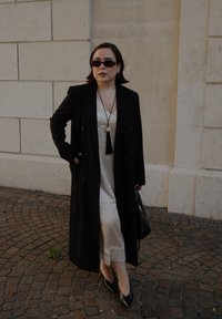Cappotto nero doppiopetto su un vestito di satin beige con pizzo. Tacchi neri a punta e una borsa nera. Occhiali da sole e una collana con nappina.