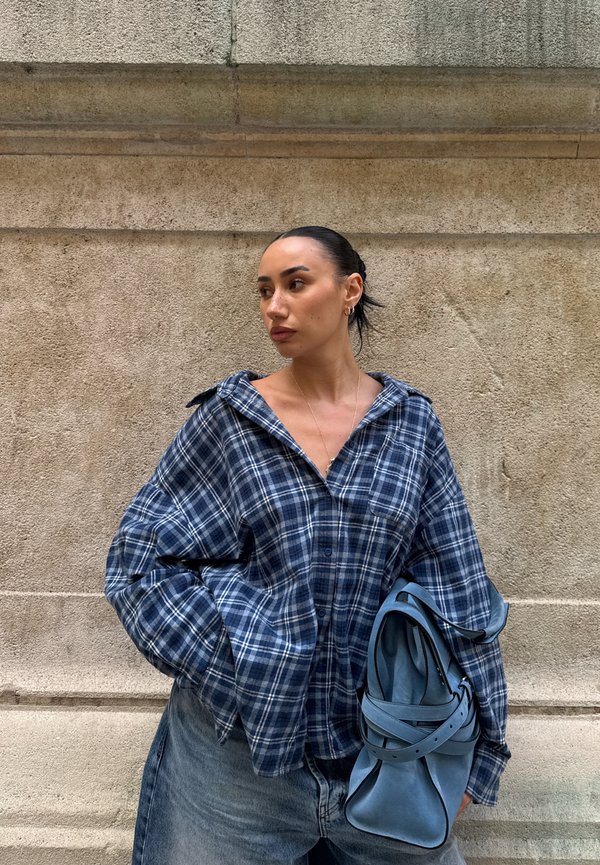 Una mujer con una camisa de cuadros azules y unos vaqueros grises está de pie junto a un muro de piedra, sostiene un bolso azul y mira hacia un lado.