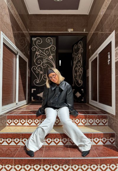 Joven con cabello rubio, gafas, chaqueta de cuero negra y pantalones blancos sentada en escalones con azulejos estampados frente a puertas metálicas decorativas.