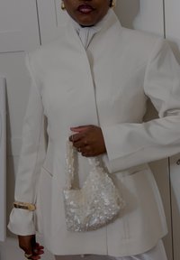 White tailored jacket with structured shoulders, a high collar, and a smooth finish. Holding a small, textured bag decorated with light sequins.
