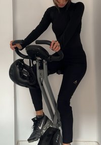 Bicicleta de exercício com um quadro preto, guiador ergonómico e um assento. O utilizador veste uma blusa preta de manga longa e leggings pretas justas, com ténis cinzentos.
