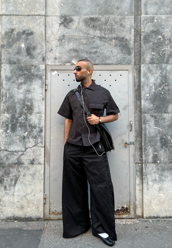Hombre con pantalones negros de pierna ancha y camisa de manga corta está junto a una puerta de metal gris, lleva gafas de sol y auriculares con un bolso negro de hombro.