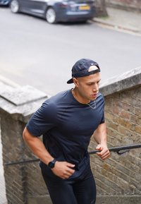 Mężczyzna ubrany w czarne odzież sportową i czapkę biegnie po zewnętrznych kamiennych schodach obok ulicy z zaparkowanymi samochodami w tle.