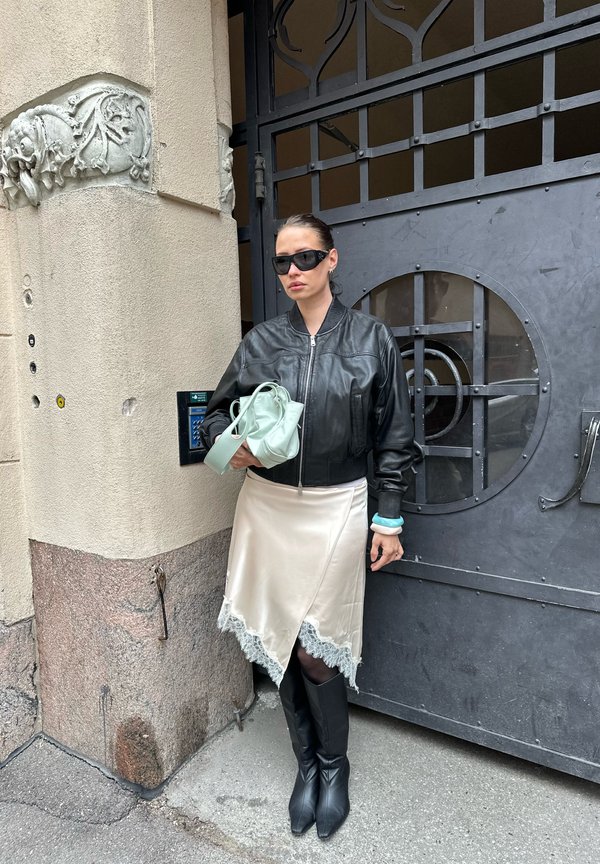 Frau mit schwarzer Lederjacke, cremefarbenem Rock mit Spitzenbesatz, schwarzen Stiefeln und Sonnenbrille steht an einer dekorativen Metalltür und hält eine minzgrüne Tasche.