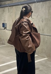 Barna műbőr kabát texturált anyaggal, laza szabással és széles ujjakkal. Hozzáillő barna kézitáskával párosítva. Beton háttér.