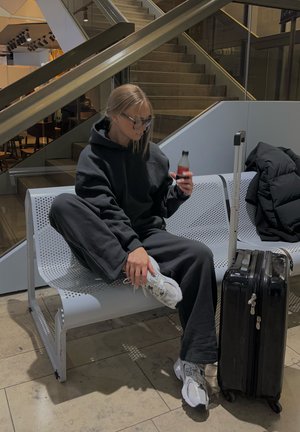 Sudadera negra con pantalones de chándal, zapatillas gris con acentos blancos. Maleta negra con herrajes plateados sobre un banco perforado en un espacio moderno.