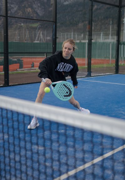 Un jugador con un sudadera negra golpea una pelota amarilla utilizando una pala de pickleball azul claro en una cancha azul. Una red está en primer plano.