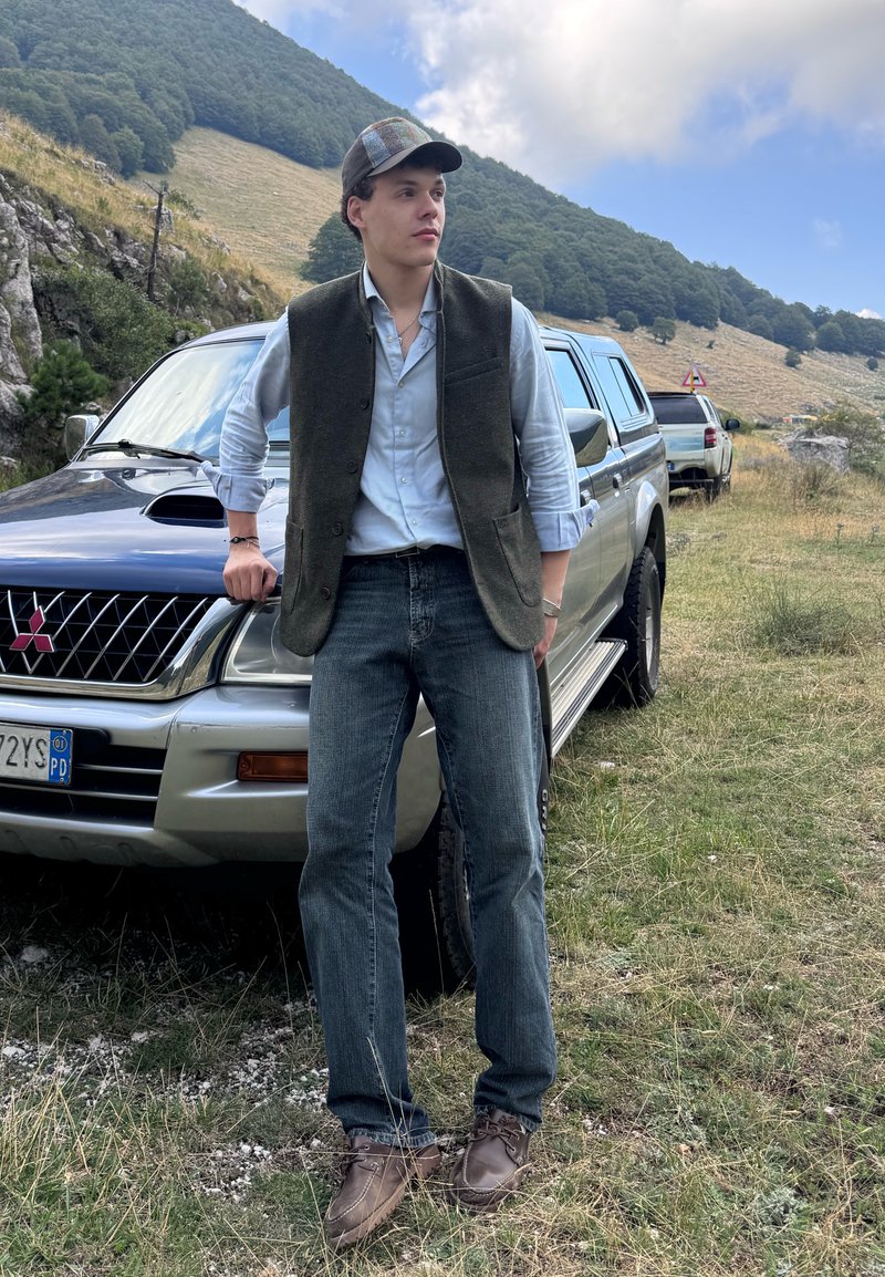 Joven con gorra, chaleco y vaqueros apoyado en una camioneta Mitsubishi plateada en una zona montañosa con hierba.