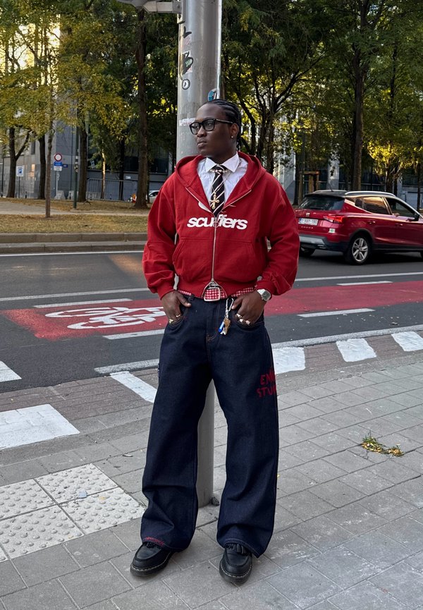 Red zip-up hoodie featuring the "scuffers" logo, a white dress shirt, a patterned tie, oversized dark denim trousers, and black shoes, standing next to a street pole.