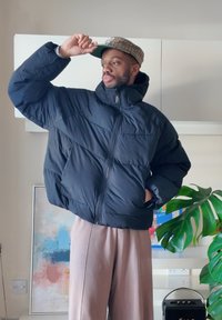 Mies, kuris dėvi juodą pūkinę striukę ir smėlio spalvos kelnes, koreguoja savo languotą skrybėlę, stovėdamas viduje šalia abstraktaus meno ir monstera augalo.