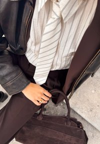 Brun mockaläder tote-väska, randig ljus skjorta i tyg, mörk jacka och bruna byxor. Närbild på hand som bär en guldring. Texturerade ytor synliga.