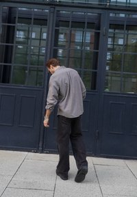 Man in een grijs shirt en een zwarte spijkerbroek loopt over een betegende grond naar donkere glazen deuren met een frame.
