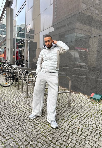 Chaqueta acolchada de color gris claro con capucha, sudadera gris y zapatillas deportivas grises. Entorno urbano con pavimento de adoquines y aparcamientos para bicicletas.