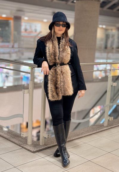 Mujer con atuendo negro, bufanda de piel sintética, gafas de sol y sombrero apoyada en una barandilla dentro de un centro comercial de varios niveles.