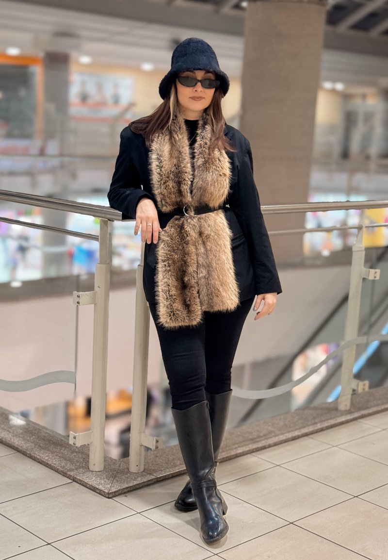 Femme en tenue noire avec écharpe en fausse fourrure, lunettes de soleil et chapeau, appuyée sur une rambarde à l'intérieur d'un centre commercial à plusieurs niveaux.