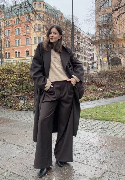 Abrigo de lana marrón sobre un suéter beige con un diseño sonriente, combinado con pantalones marrones de pierna ancha y zapatos negros puntiagudos. Entorno urbano.