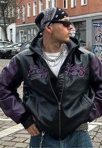 Veste en cuir noir et violet avec des accents en chaîne argentée, comportant un texte brodé, une fermeture éclair et une capuche. Portée avec un jean bleu.