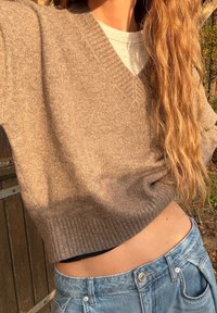 Beżowy sweter z dzianiny z dekoltem w serek i prążkowanym brzegiem, noszony na szarej koszulce i zestawiony z jasnoniebieskimi szortami dżinsowymi.