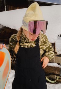 Jeune femme dans un paysage enneigé ajustant un pantalon de neige noir, portant un bonnet en tricot beige, une polaire verte à motifs, des lunettes de ski roses, tenant un snowboard.