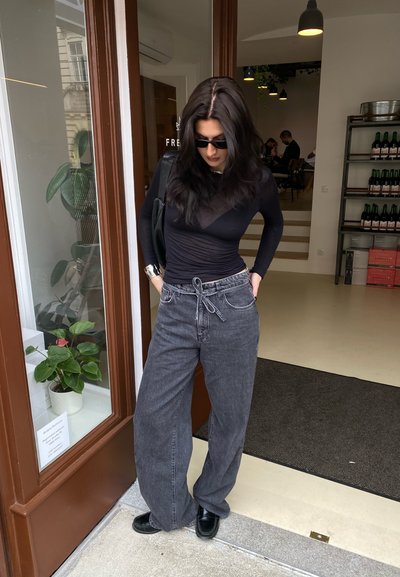 Top negro transparente de manga larga, jeans gris de tiro alto de pierna ancha con cinturón de lazo, zapatos negros tipo deslizamiento, pulsera de plata en la muñeca, ambiente interior.