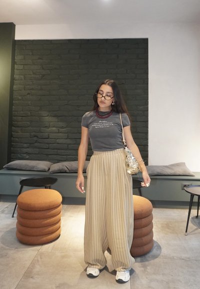 Camiseta gris con texto, pantalones de pierna ancha a rayas beige, gafas redondas y un bolso de lentejuelas. El fondo presenta una pared verde oscura y cojines.