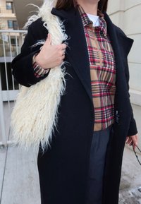 Personne portant un manteau noir et une chemise à carreaux, tenant un sac à main blanc et duveteux à l'épaule, et tenant des lunettes de soleil à la main.