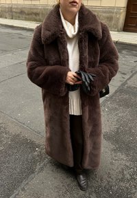 Άτομο που φοράει ένα μακρύ καφέ παλτό από συνθετική γούνα πάνω από ένα λευκό πουλόβερ, κρατώντας μαύρα δερμάτινα γάντια και μια μαύρη τσάντα χεριού με χρυσό λογότυπο.