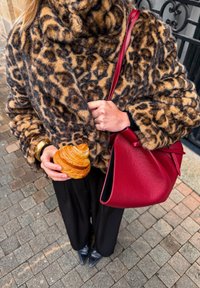 Manteau en fausse fourrure imprimé léopard, pantalon noir, sac à main en cuir rouge, tenant un croissant feuilleté. Apparence texturée, douce et chaleureuse.
