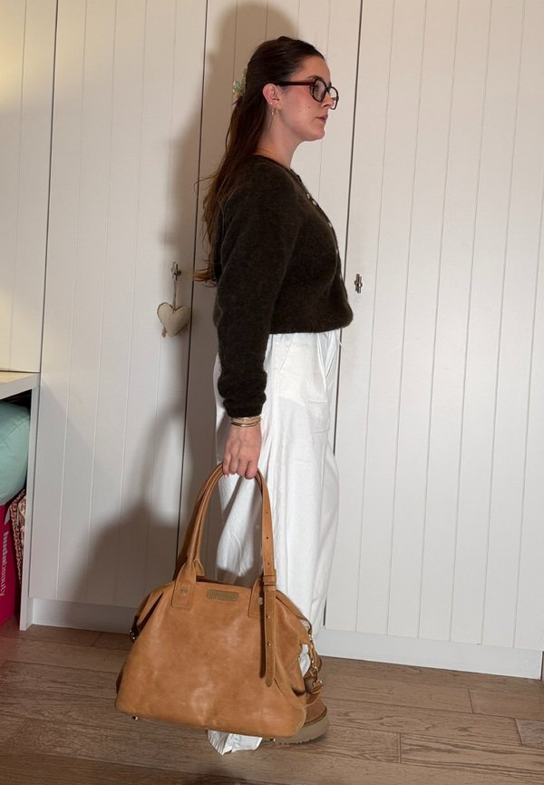 Femme aux cheveux longs et lunettes, vue de profil, portant un pull sombre et un pantalon blanc, tenant un grand sac à main en cuir marron.
