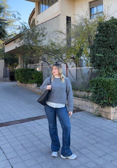 Suéter gris de punto sobre una camisa blanca con cuello, vaqueros acampanados azules y zapatillas grises, sosteniendo una bolsa tote negra al aire libre.