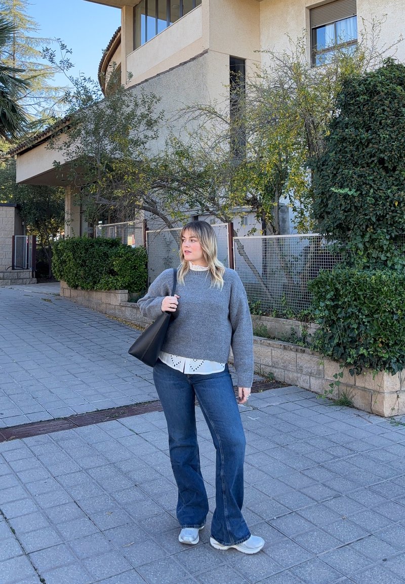 Camisola cinzenta de malha sobre camisa branca com colarinho, jeans azuis flare e ténis cinzentos, segurando uma mala preta ao ar livre.
