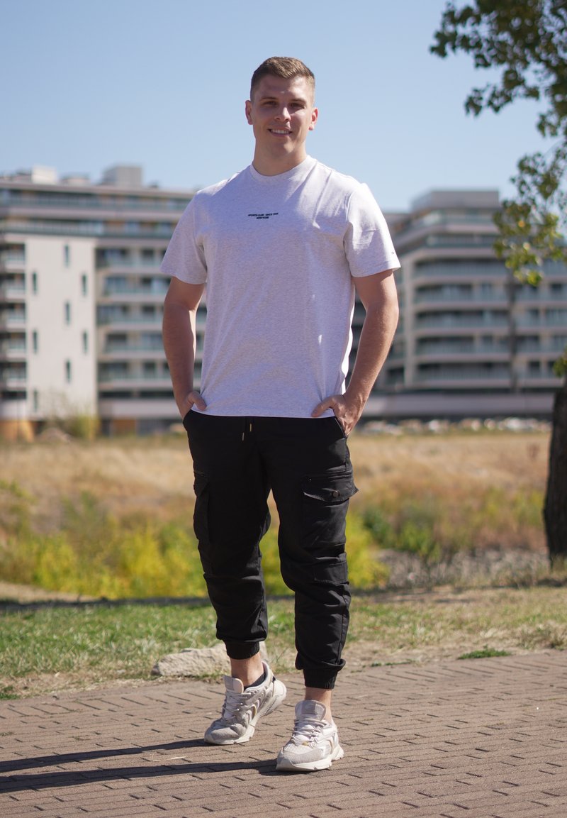 Jeune homme debout à l'extérieur sur un chemin pavé, portant un t-shirt blanc, un pantalon cargo noir et des baskets blanches, avec des bâtiments en arrière-plan.
