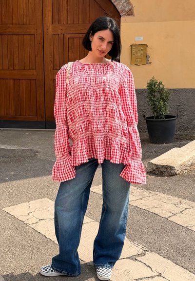 Mujer con corte de pelo bob negro que lleva una blusa a cuadros rojos y vaqueros azules holgados, de pie sobre un paso de peatones agrietado cerca de una gran puerta de madera.