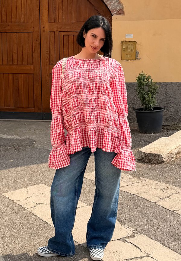 Femme avec une coupe au carré noire portant un chemisier à carreaux rouges et un jean bleu ample, se tient sur un passage piéton fissuré près d'une grande porte en bois.