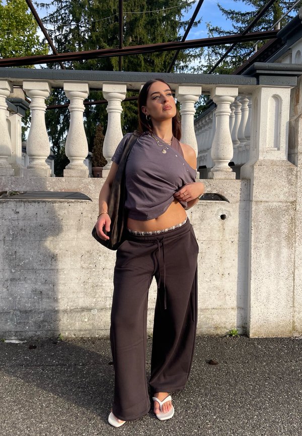 Joven mujer de pie al aire libre junto a una balaustrada de piedra blanca, vistiendo pantalones anchos oscuros, un top gris corto, sandalias blancas y llevando una bolsa negra.