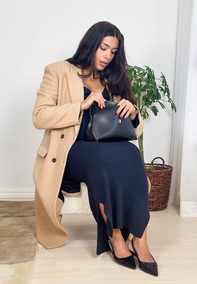 Mujer con abrigo camel y vestido negro sentada en una silla, mirando dentro de un bolso negro, con una planta en maceta al fondo.