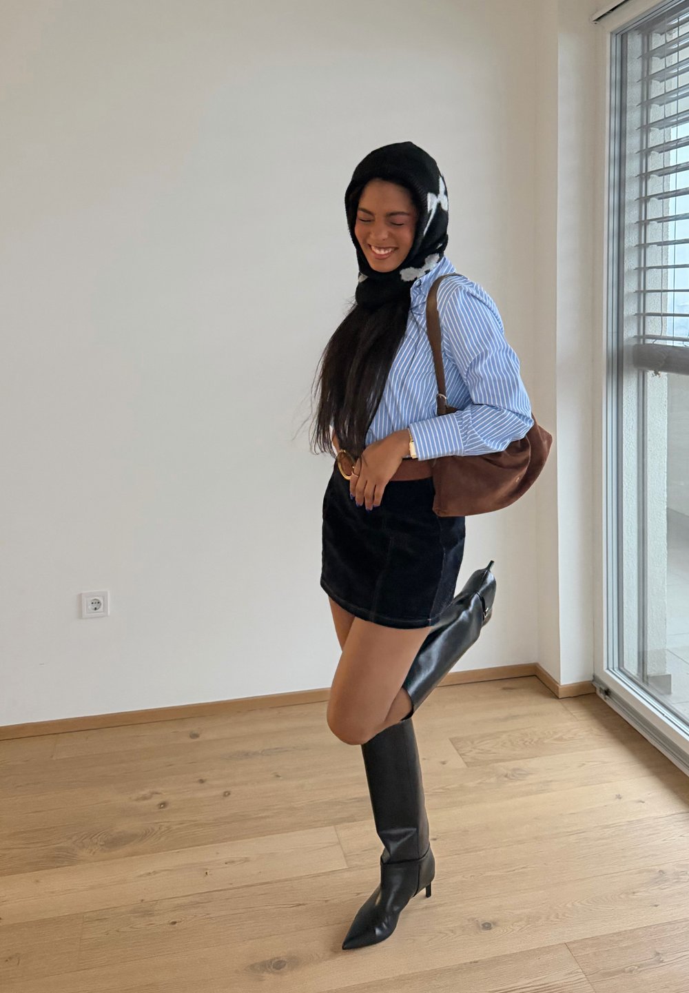 Femme portant un foulard noir et blanc, une chemise rayée bleue, une jupe noire et de hautes bottes noires, souriant en se tenant près d'une fenêtre.