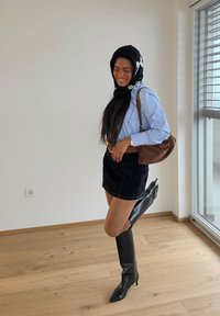 Femme portant un foulard noir et blanc, une chemise rayée bleue, une jupe noire et de hautes bottes noires, souriant en se tenant près d'une fenêtre.